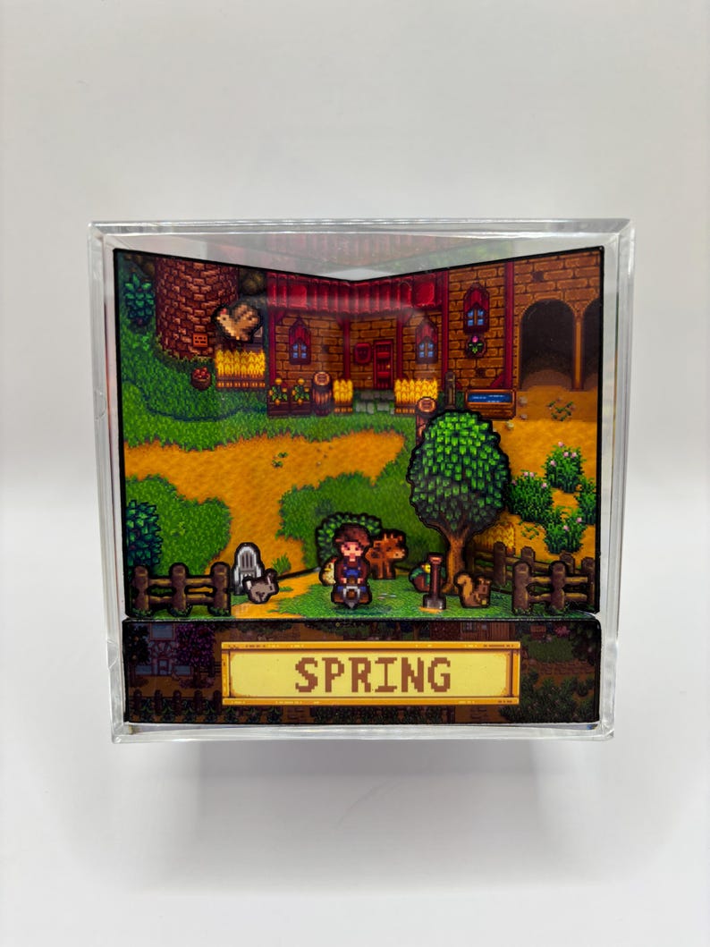 May include: A diorama featuring a pixelated scene with a character, animals, and a building, all within a clear acrylic cube. The word "SPRING" is displayed on a golden banner at the bottom. The scene is set in a vibrant, colorful environment.