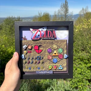 Op de afbeelding: Een ingelijst kunstwerk met de interface van het spel "Legend of Zelda: Ocarina of Time". Het kunstwerk bevat het logo van het spel, de queststatus, hartcontainers en muzieknoten. De lijst is zwart, met een landschappelijke achtergrond.