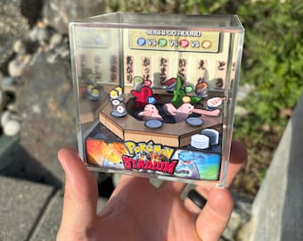 May include: A clear acrylic cube featuring a diorama of a Pokemon Stadium scene. The diorama depicts a sushi-themed battle arena with various Pokemon characters and sushi elements. The text "SUSHI GO-ROUND" and "Pokemon Stadium" are visible.
