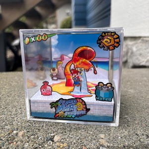 May include: A clear acrylic display box featuring a Super Mario Sunshine diorama. The scene includes Mario, a water spout, and various game elements. The box has a printed base with the game's logo and a tropical theme.