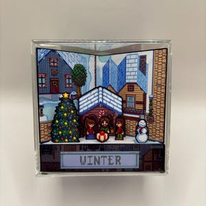 May include: A clear acrylic block featuring a pixelated winter scene. The scene includes a decorated Christmas tree, snow-covered houses, and three figures. The word "WINTER" is displayed at the bottom of the block.