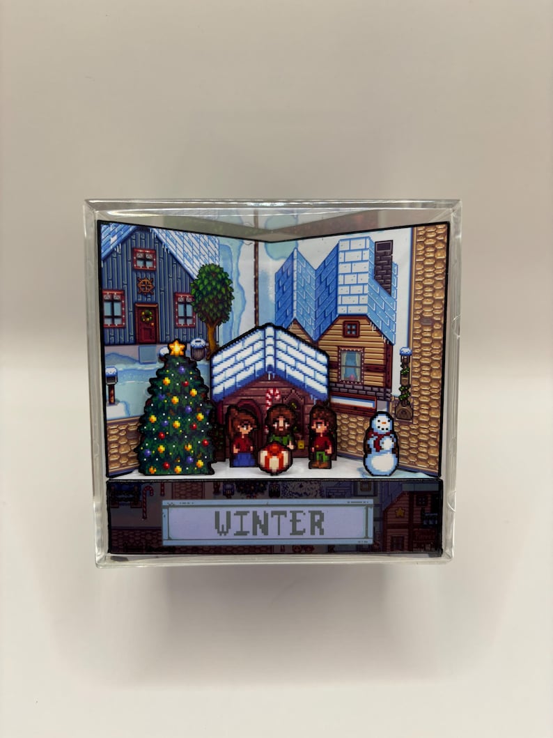 May include: A clear acrylic block featuring a pixelated winter scene. The scene includes a decorated Christmas tree, snow-covered houses, and three figures. The word "WINTER" is displayed at the bottom of the block.