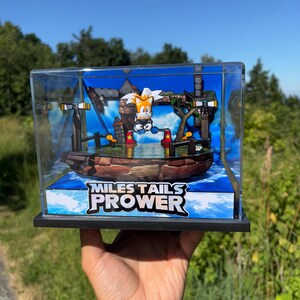 Op de afbeelding: Een doorzichtige display case met een diorama van het personage Miles "Tails" Prower uit de Sonic the Hedgehog-serie. Het diorama bevat een boot, water en de tekst "Miles Tails Prower". De achtergrond is een blauwe lucht.