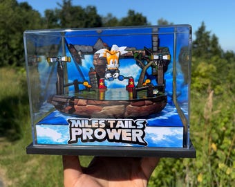 May include: A clear display case containing a diorama of the character Miles "Tails" Prower from the Sonic the Hedgehog series. The diorama features a boat, water, and the text "Miles Tails Prower". The background is a blue sky.