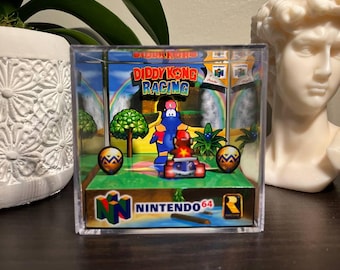 May include: A clear acrylic display case featuring a miniature diorama of the Nintendo 64 game, Diddy Kong Racing. The scene includes Diddy Kong in a kart, a rainbow, and the Nintendo 64 logo. The case is on a dark surface.