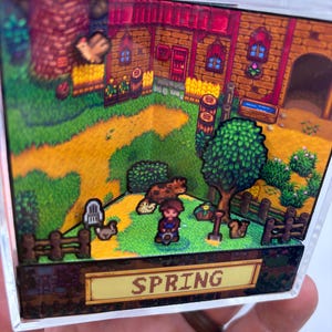 May include: A 3D diorama depicting a farm scene with a house, trees, and a dirt path. The word "SPRING" is displayed on a yellow sign. The scene includes a character, animals, and a fence, all within a clear, rectangular frame.