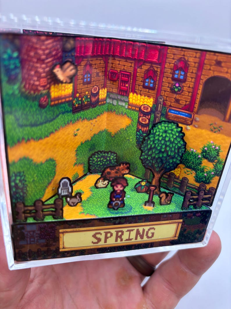 May include: A 3D diorama depicting a farm scene with a house, trees, and a dirt path. The word "SPRING" is displayed on a yellow sign. The scene includes a character, animals, and a fence, all within a clear, rectangular frame.