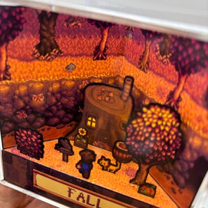 May include: A miniature diorama scene with a fall theme. The scene includes a tree stump house, trees with orange and red leaves, and the word "FALL" in a banner. A small figure and a cat are also present.
