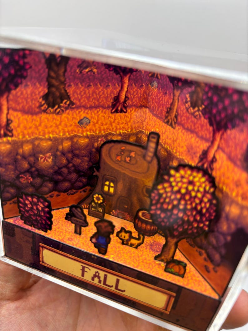 May include: A miniature diorama scene with a fall theme. The scene includes a tree stump house, trees with orange and red leaves, and the word "FALL" in a banner. A small figure and a cat are also present.