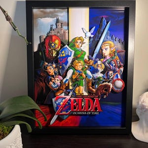 Op de afbeelding: Ingelijste kunstwerk met personages uit de videogame "The Legend of Zelda: Ocarina of Time". Het kunstwerk heeft een 3D-effect, met gelaagde personages en elementen in een zwart frame. De titel wordt weergegeven in rode en witte tekst.
