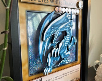 May include: Framed artwork featuring a 3D rendering of the "Blue-Eyes White Dragon" card from the Yu-Gi-Oh! trading card game. The dragon is depicted in shades of blue and white, set against a textured background. The frame is black.
