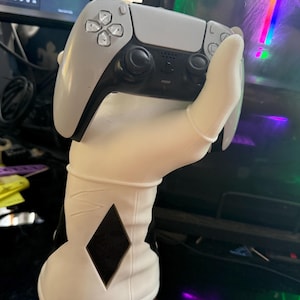 Peut inclure: Un support de manette de jeu blanc et noir en forme de main, tenant une manette grise et noire. Le support présente une découpe en forme de losange. La manette est une PlayStation 5 DualSense.