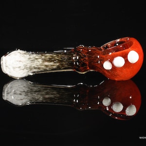 May include: A red glass pipe with white polka dots. The pipe is made of glass and has a long, thin stem. The bowl of the pipe is round and has a small hole in the center.