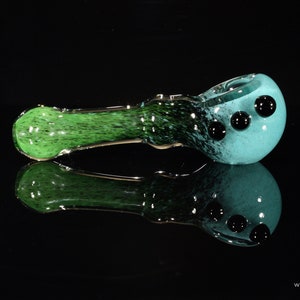 May include: A green and blue glass pipe with black dots. The pipe has a rounded bowl and a long, thin stem.