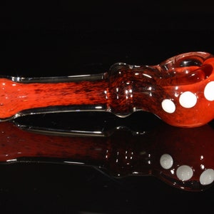 May include: A red glass pipe with white polka dots. The pipe has a long, thin stem and a bowl with a rounded shape.