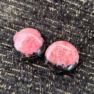 May include: Two round, polished stones with a pink center and a black border. The stones are set against a dark, textured background. The pink color is a vibrant contrast to the black.