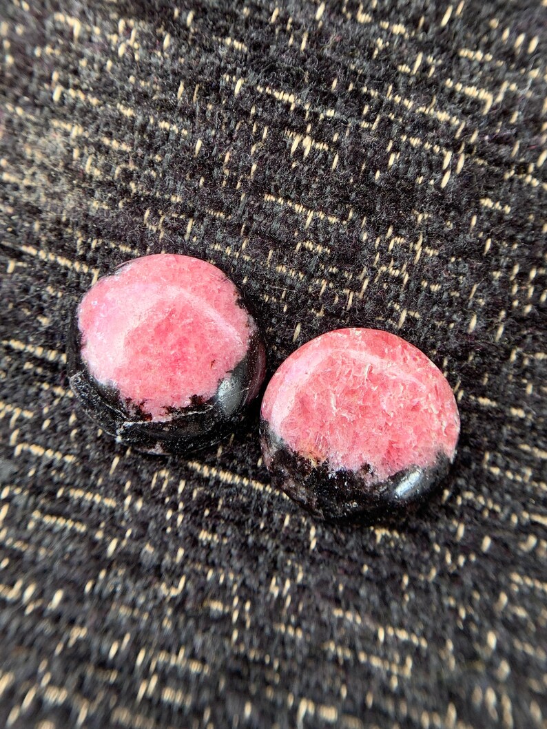 May include: Two round, polished stones with a pink center and a black border. The stones are set against a dark, textured background. The pink color is a vibrant contrast to the black.
