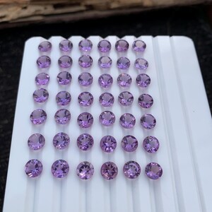 May include: A collection of round, faceted amethyst gemstones in varying shades of purple. The gemstones are arranged on a white tray, showcasing their clarity and color. The gemstones are ready for jewelry making.