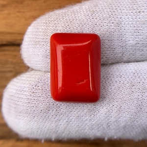 May include: A rectangular, polished, vibrant red gemstone. The stone has rounded edges and a smooth, glossy surface. The gemstone is held against a white fabric background.