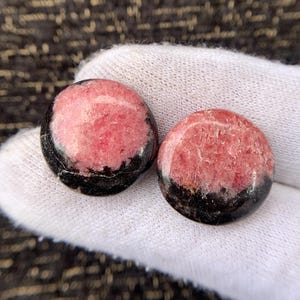 May include: Two round, polished stones. Each stone has a pink center with a black border. The stones are displayed on a white surface, with a dark, textured background.