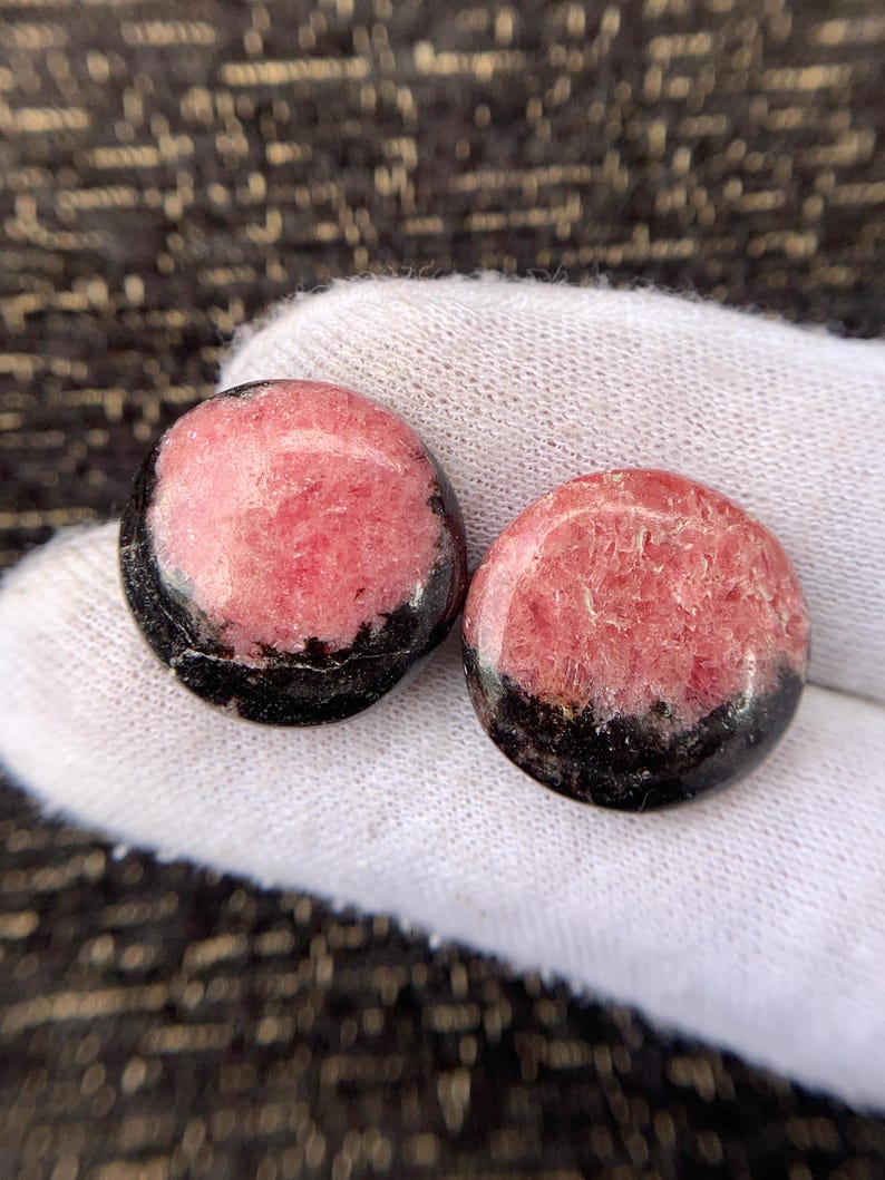 May include: Two round, polished stones. Each stone has a pink center with a black border. The stones are displayed on a white surface, with a dark, textured background.