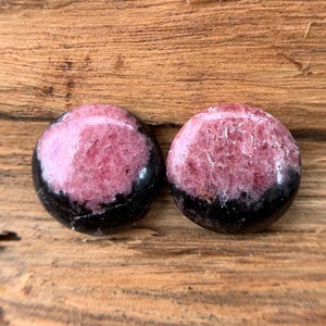 May include: Two round, polished gemstones. Each stone features a vibrant pink center with a black band around the bottom. The stones are displayed on a textured, wooden surface, showcasing their natural beauty and color variations.