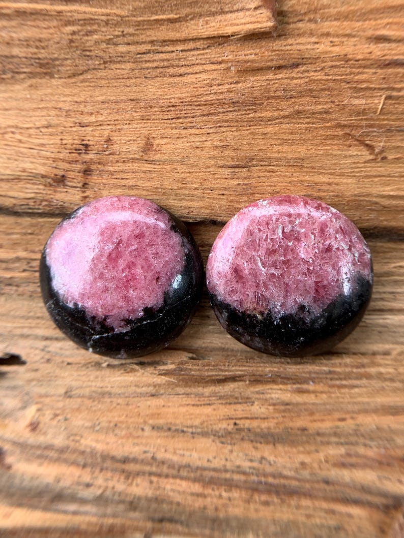 May include: Two round, polished gemstones. Each stone features a vibrant pink center with a black band around the bottom. The stones are displayed on a textured, wooden surface, showcasing their natural beauty and color variations.