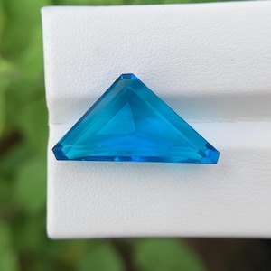 Pode incluir: Uma pedra preciosa triangular e facetada em um tom vibrante de azul. A gema está colocada sobre uma superfície branca, com um fundo verde desfocado. O corte da gema permite que a luz se reflita, criando um efeito brilhante. Esta gema pode ser usada em joias.