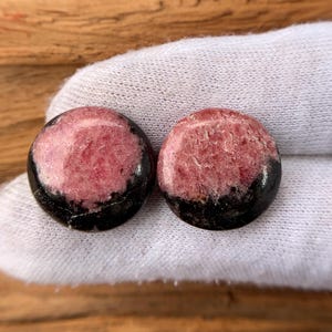 May include: Two round, polished stones. Each stone has a pink center with a black base. The stones are displayed on a white surface, with a wooden background.