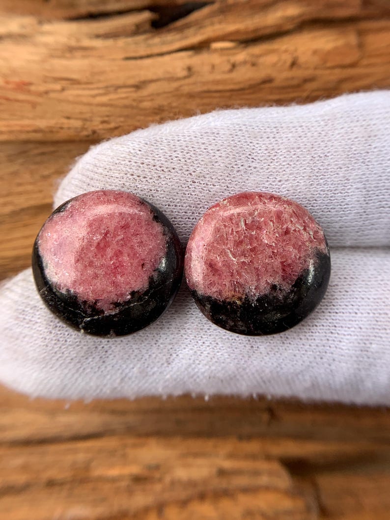 May include: Two round, polished stones. Each stone has a pink center with a black base. The stones are displayed on a white surface, with a wooden background.