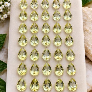 May include: A collection of thirty-five faceted, pear-shaped lemon quartz gemstones arranged in a grid on a rectangular beige display pad. Each translucent, pale yellow stone features a pointed bottom and a rounded top with multiple facets.