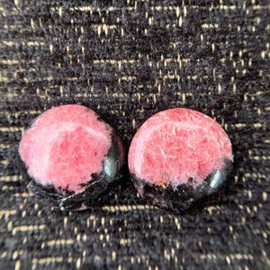May include: Two polished, round gemstones. Each stone features a vibrant pink center with a band of black along the bottom edge. The stones are set against a textured, dark background.