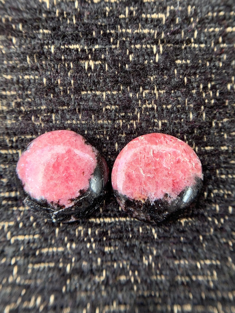 May include: Two polished, round gemstones. Each stone features a vibrant pink center with a band of black along the bottom edge. The stones are set against a textured, dark background.