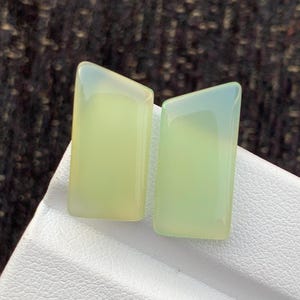 May include: Two smooth, polished, light green rectangular gemstones with slightly angled top edges. These translucent stones feature a soft, pale lime hue and a glossy, reflective surface, showcasing a uniform colour and a clean, flat, geometric shape.