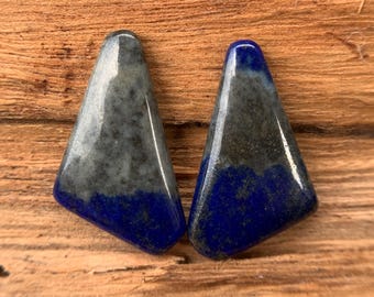May include: Two polished, triangular gemstones. The top portion of each stone is a speckled gray, transitioning to a deep blue at the base. The stones are displayed on a wooden surface.