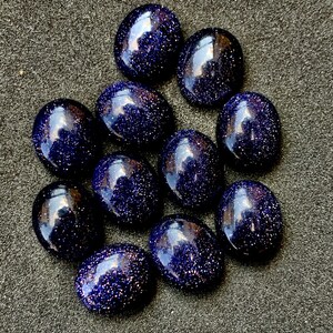 May include: A collection of eleven polished, oval-shaped blue sandstone gemstones. The stones have a deep blue color with sparkling inclusions, giving them a starry appearance. They are arranged on a dark gray surface.