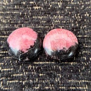May include: Two round, polished stones with a pink and black color scheme. The pink portion is a lighter shade with darker veining, while the bottom half is a solid black. The stones are set against a textured, dark background.