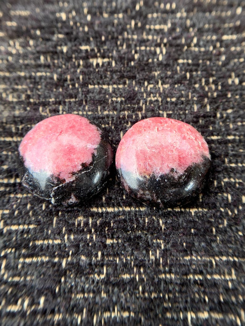 May include: Two round, polished stones with a pink and black color scheme. The pink portion is a lighter shade with darker veining, while the bottom half is a solid black. The stones are set against a textured, dark background.