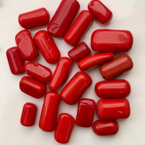 May include: A selection of polished, vivid red coral beads. The beads are cylindrical and rectangular, with rounded edges. The coral pieces vary in size and are scattered on a white surface. The colour is a deep, saturated red.