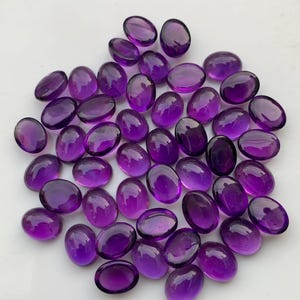 May include: A collection of oval-shaped, polished amethyst gemstones. The gemstones are a deep purple color and have a smooth, glossy surface. They are scattered on a white surface, creating a visually appealing display.