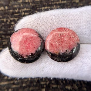 May include: Two round gemstones with a pink and black color scheme. The pink portion has a textured appearance, while the bottom is a solid black. The gemstones are displayed on a white surface.