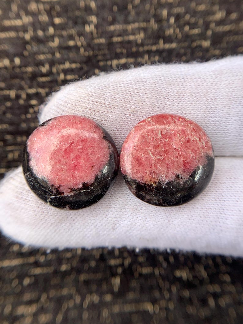 May include: Two round gemstones with a pink and black color scheme. The pink portion has a textured appearance, while the bottom is a solid black. The gemstones are displayed on a white surface.