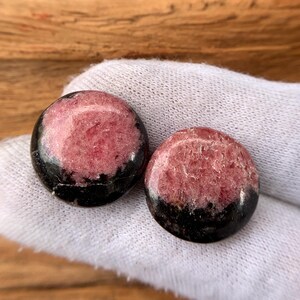 May include: Two round, polished gemstones. Each stone features a pinkish-red center with a black band around the bottom edge. The stones are held on a white fabric, with a wooden background.