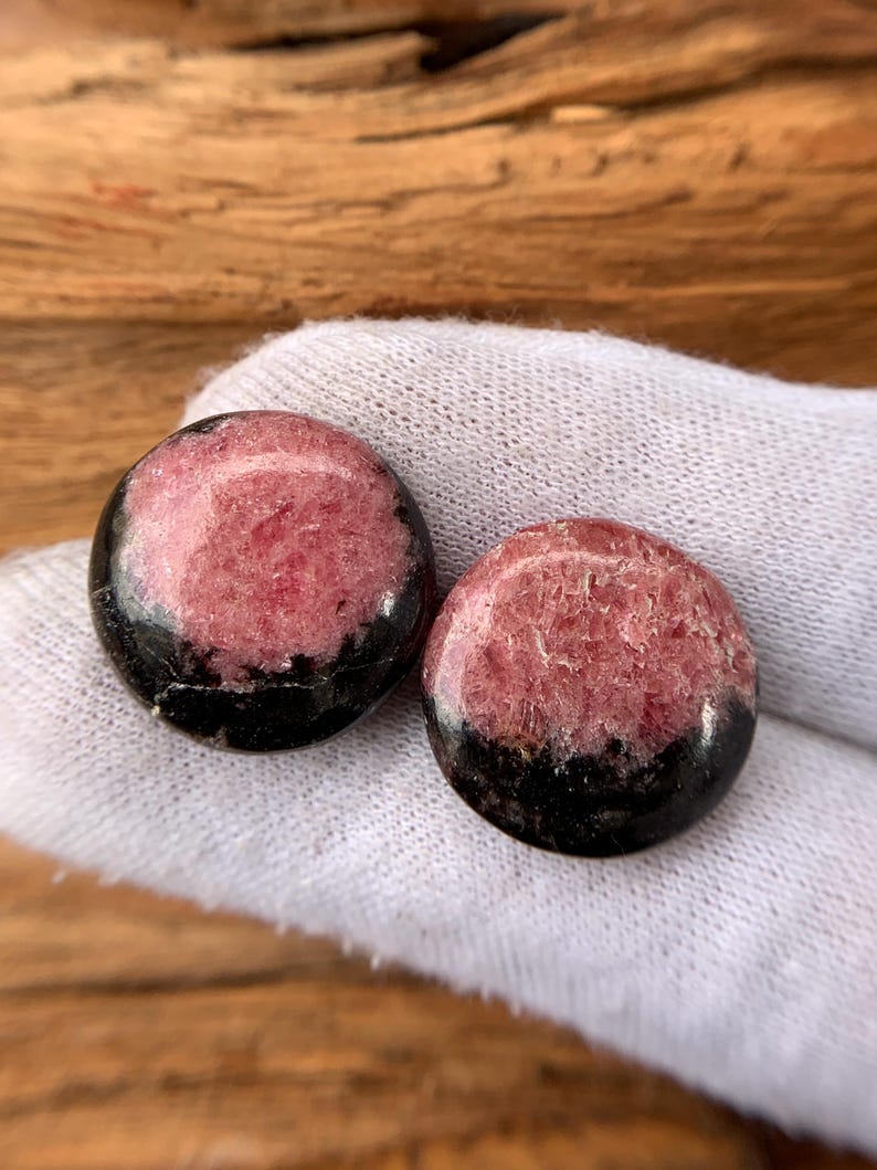May include: Two round, polished gemstones. Each stone features a pinkish-red center with a black band around the bottom edge. The stones are held on a white fabric, with a wooden background.