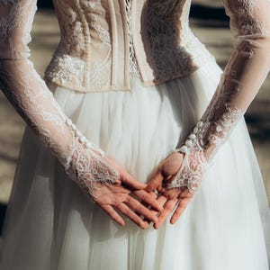 Könnte beinhalten: Ein cremefarbenes Hochzeitskleid mit einem Korsett mit Spitzendetails und einem weißen Tüllrock. Die langen Ärmel sind aus zarter Spitze gefertigt, und die Hände sind vor der Brust gefaltet. Das Kleid ist elegant.