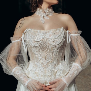 Könnte beinhalten: Weißes Brautkleid mit aufwendigen Spitzendetails. Das Kleid hat einen herzförmigen Ausschnitt, transparente Ärmel und einen passenden Spitzen-Choker. Das Oberteil ist mit floralen und perlenbesetzten Verzierungen versehen, die einen romantischen und eleganten Look erzeugen.