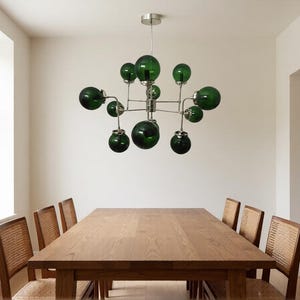 Op de afbeelding: Een moderne eetkamer met een grote houten tafel en acht stoelen met geweven rugleuningen. Boven de tafel hangt een unieke kroonluchter met meerdere donkergroene glazen bolvormige lampenkappen en een zilverkleurig metalen frame.
