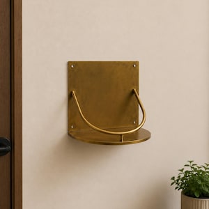 Puede incluir: Un estante de pared hecho de metal de latón cepillado. Cuenta con una placa trasera cuadrada con cuatro orificios para tornillos en las esquinas, una base semicircular y una barra de soporte curva en forma de U que une la placa con el borde.