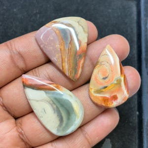 May include: Three polished gemstones in shades of beige, pink, orange, and green. The stones have unique patterns and shapes, including a triangle and two teardrop forms. The gemstones are held in a hand, showcasing their natural beauty and intricate designs.