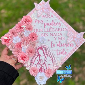 May include: A decorated graduation cap with pink paper flowers, butterflies, and text in Spanish. The cap features a central image of a religious figure and the words "PARA mis padres QUE LLEGARON SIN NADA Y ME lo dieron todo."